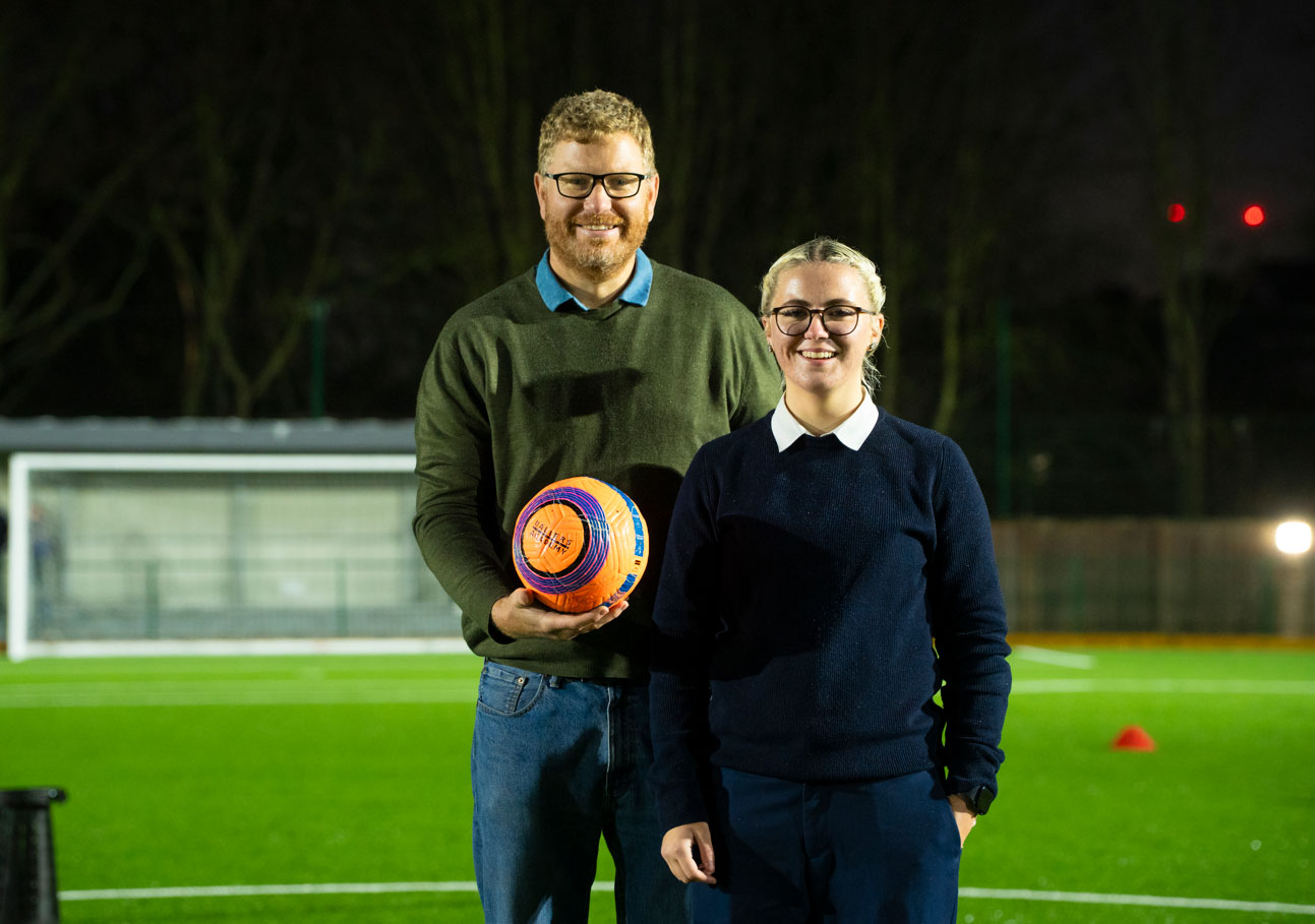 Abigail and Pete standing on an outdoor football pitch holding a football.