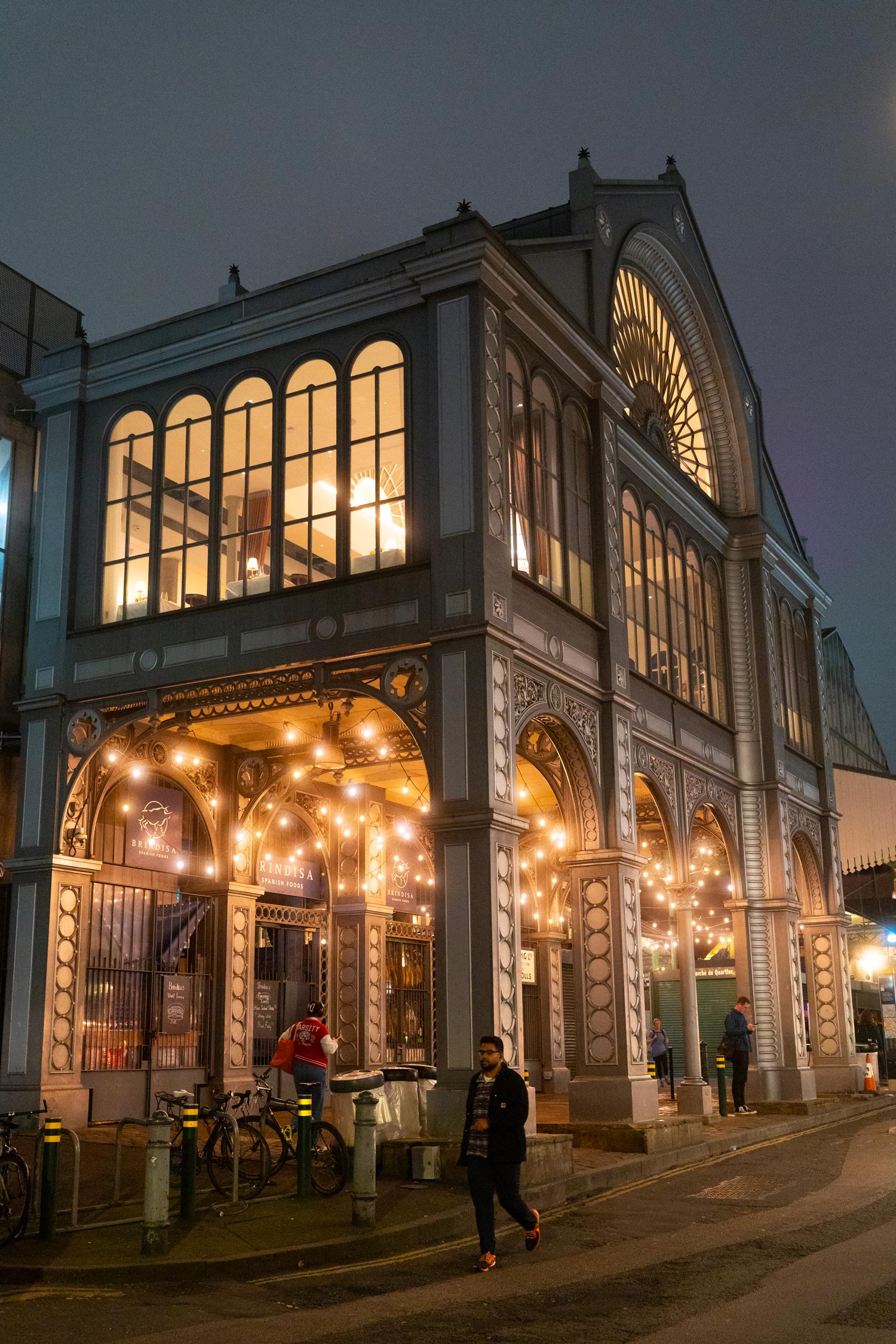 Exterior of Borough Market at night with warm lights illuminating the historic market building and people walking nearby.